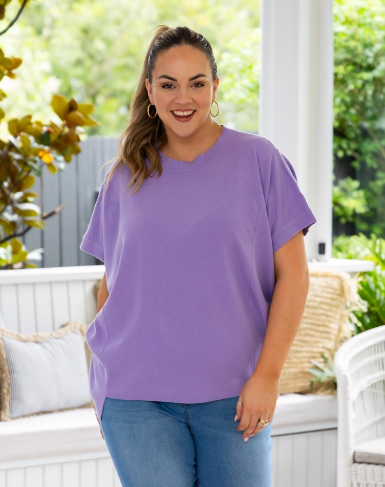 A woman modeling a loose-fit, short-sleeve purple t-shirt with blue jeans, standing on a white porch with green foliage in the background.