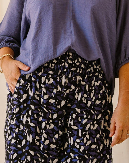 A close-up of a person wearing loose black pants with a blue and white abstract leaf pattern, paired with a lavender blouse.