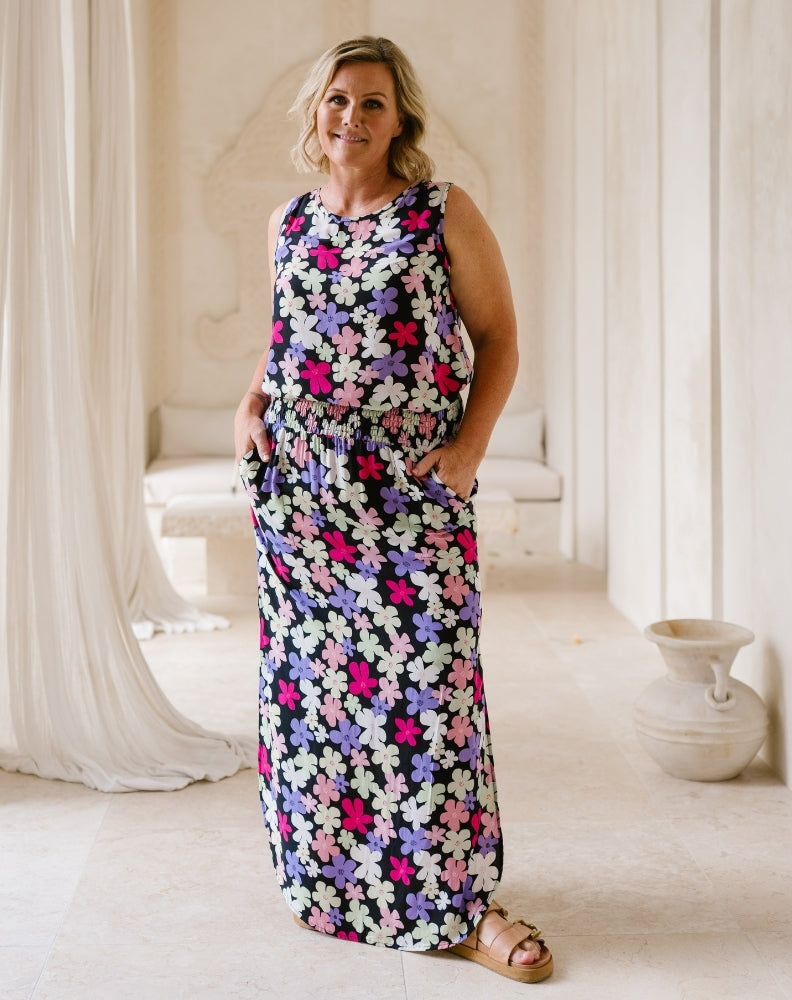A woman modeling a sleeveless, floor-length maxi dress with a colorful floral pattern on a dark background, standing in a bright, neutral-toned indoor setting.