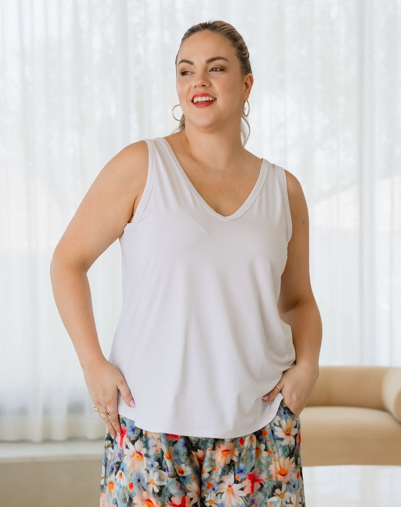 Woman wearing a white tank top and colorful floral pants against a white curtain background