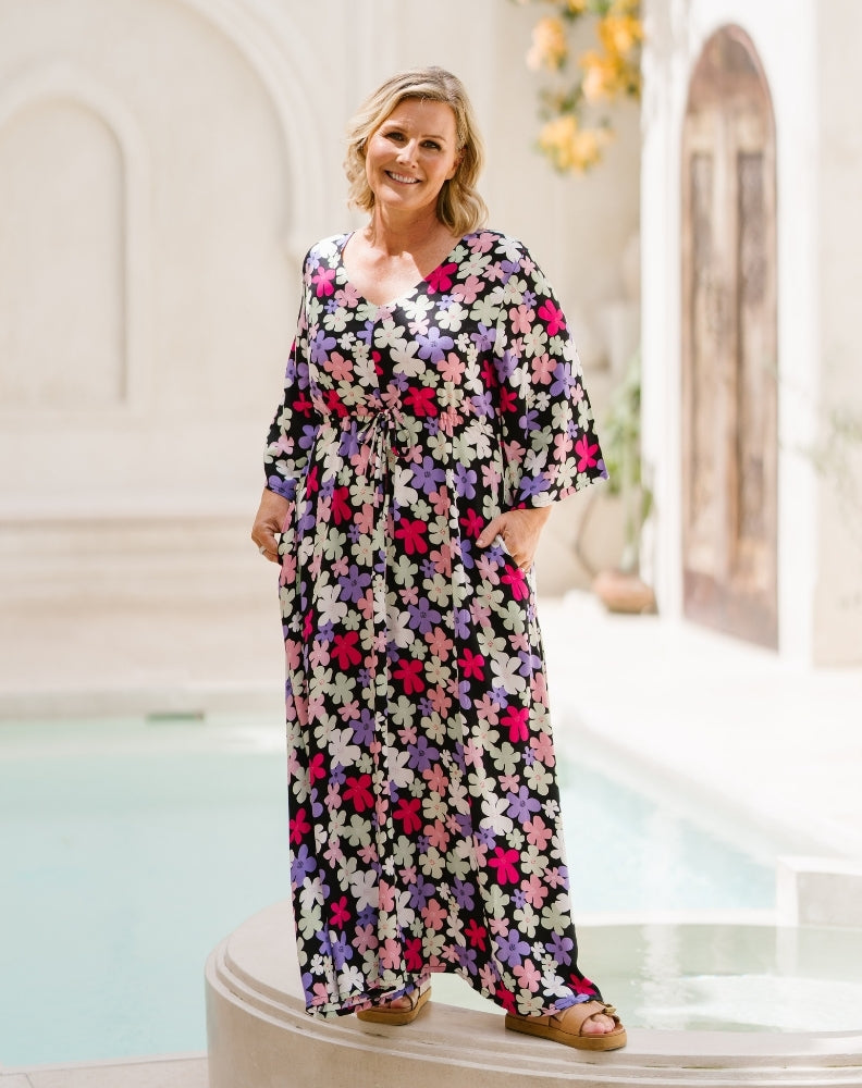 A woman wearing a long-sleeve maxi dress with a colorful floral print stands on a white architectural platform next to a pool.