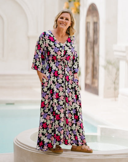 A woman wearing a black maxi dress with a colorful floral pattern of pink, purple, and white flowers, standing on a white architectural platform next to a pool.