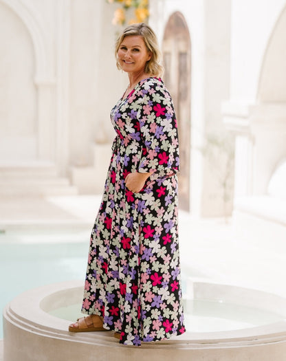 A woman wearing a long-sleeved, knee-length maxi dress with a vibrant floral print in pink, purple, white, and black, standing on a white stone terrace in a bright, sunlit architectural setting.