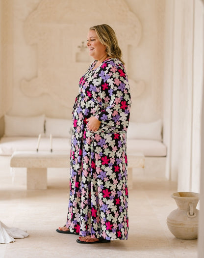 A woman wearing a long-sleeve maxi dress with a colorful floral pattern, standing in a bright, neutral-toned indoor setting with a sofa and decorative vase in the background.