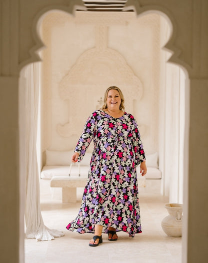 A woman wearing a long-sleeved maxi dress with a colorful floral pattern stands in a bright, cream-colored architectural setting framed by arches.