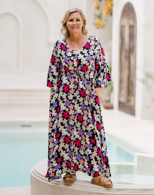 A woman wearing a long-sleeve, V-neck maxi dress with a colorful floral print, standing in front of a white architectural background with a pool.