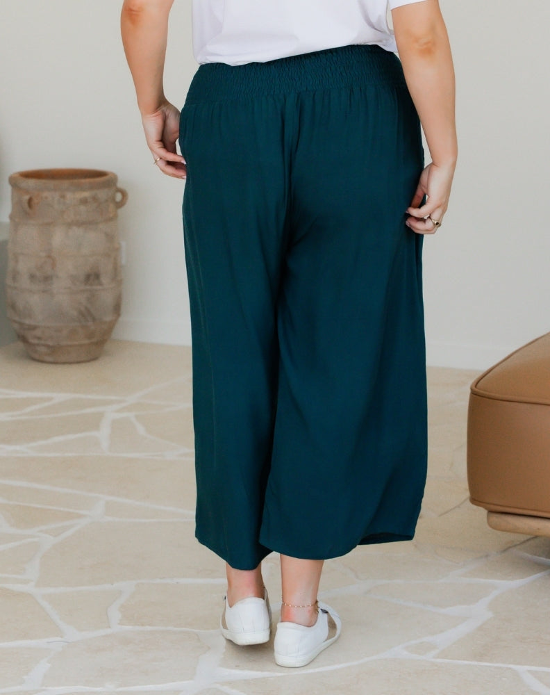 Person wearing teal wide-leg pants with a white top and white shoes, standing in a room with a vase and chair.